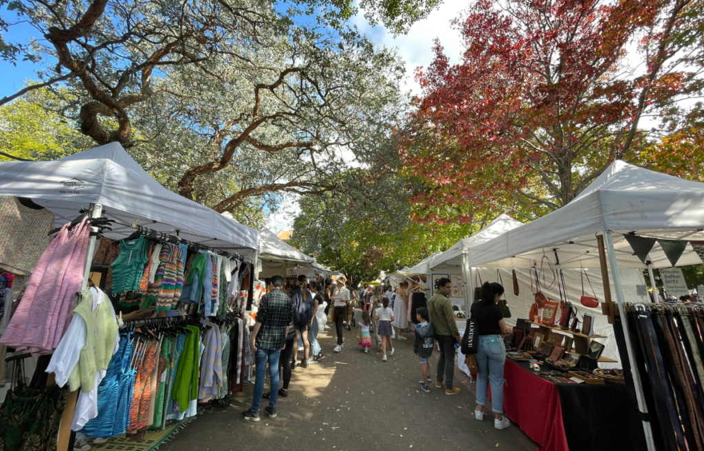 Glebe Market in Sydney, Australia, with people browsing stalls under white tents selling colorful clothing and leather goods. Large trees with green and red leaves provide shade.